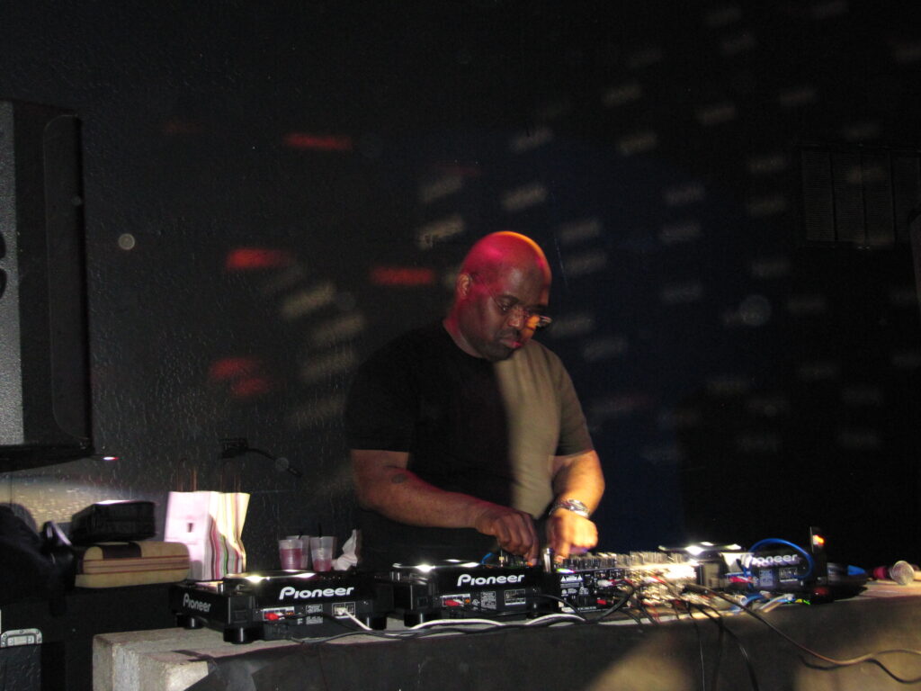Frankie Knuckles concentré derrière une console DJ dans un club sombre éclairé de rouge.