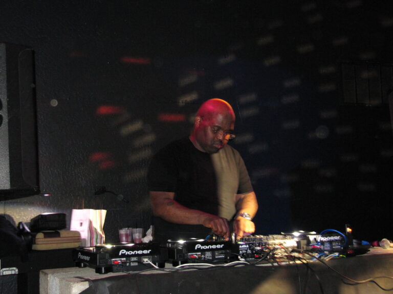 Frankie Knuckles concentré derrière une console DJ dans un club sombre éclairé de rouge.