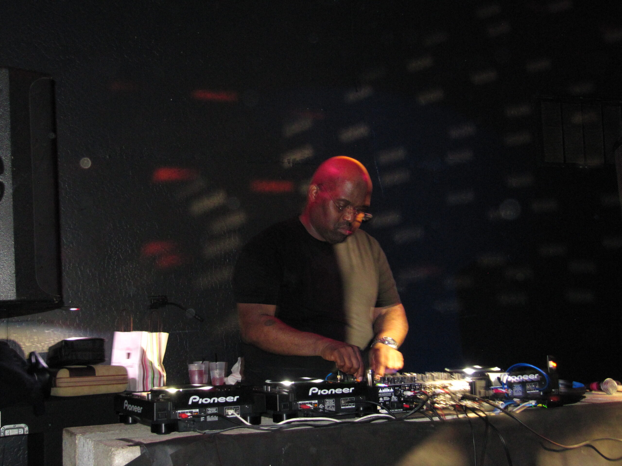 Frankie Knuckles concentré derrière une console DJ dans un club sombre éclairé de rouge.